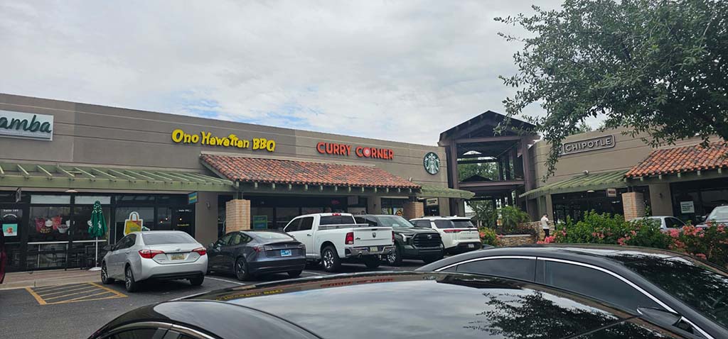 A shopping plaza featuring Curry Corner, Starbucks, and Chipotle restaurants, with parked cars in front.